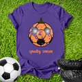 A purple short-sleeve t-shirt laid flat on green grass, featuring a graphic of a soccer ball turned into a pumpkin with the words 'spooky season' below it. A real soccer ball and black cleats are placed around the shirt as props.