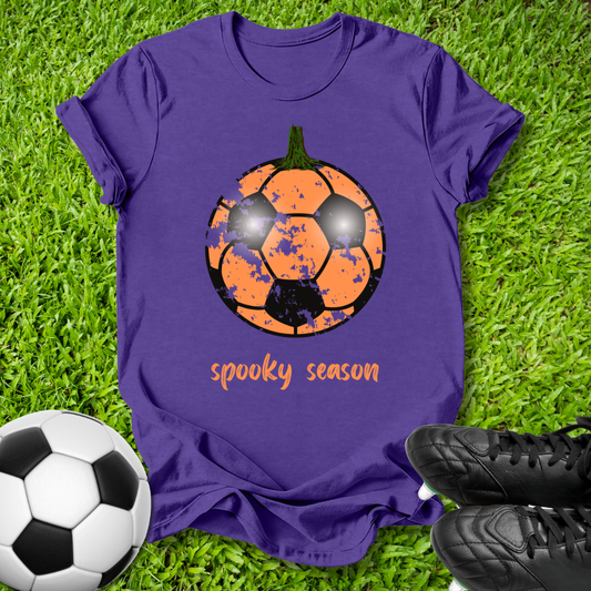 A purple short-sleeve t-shirt laid flat on green grass, featuring a graphic of a soccer ball turned into a pumpkin with the words 'spooky season' below it. A real soccer ball and black cleats are placed around the shirt as props.