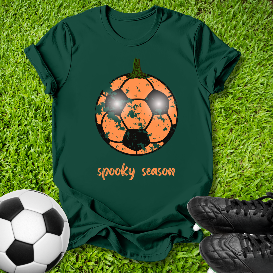 A dark green short-sleeve t-shirt laid flat on green grass, featuring a distressed orange soccer pumpkin graphic with the words 'spooky season' in orange script below it. A black-and-white soccer ball is at the bottom left, and black cleats are at the bottom right.