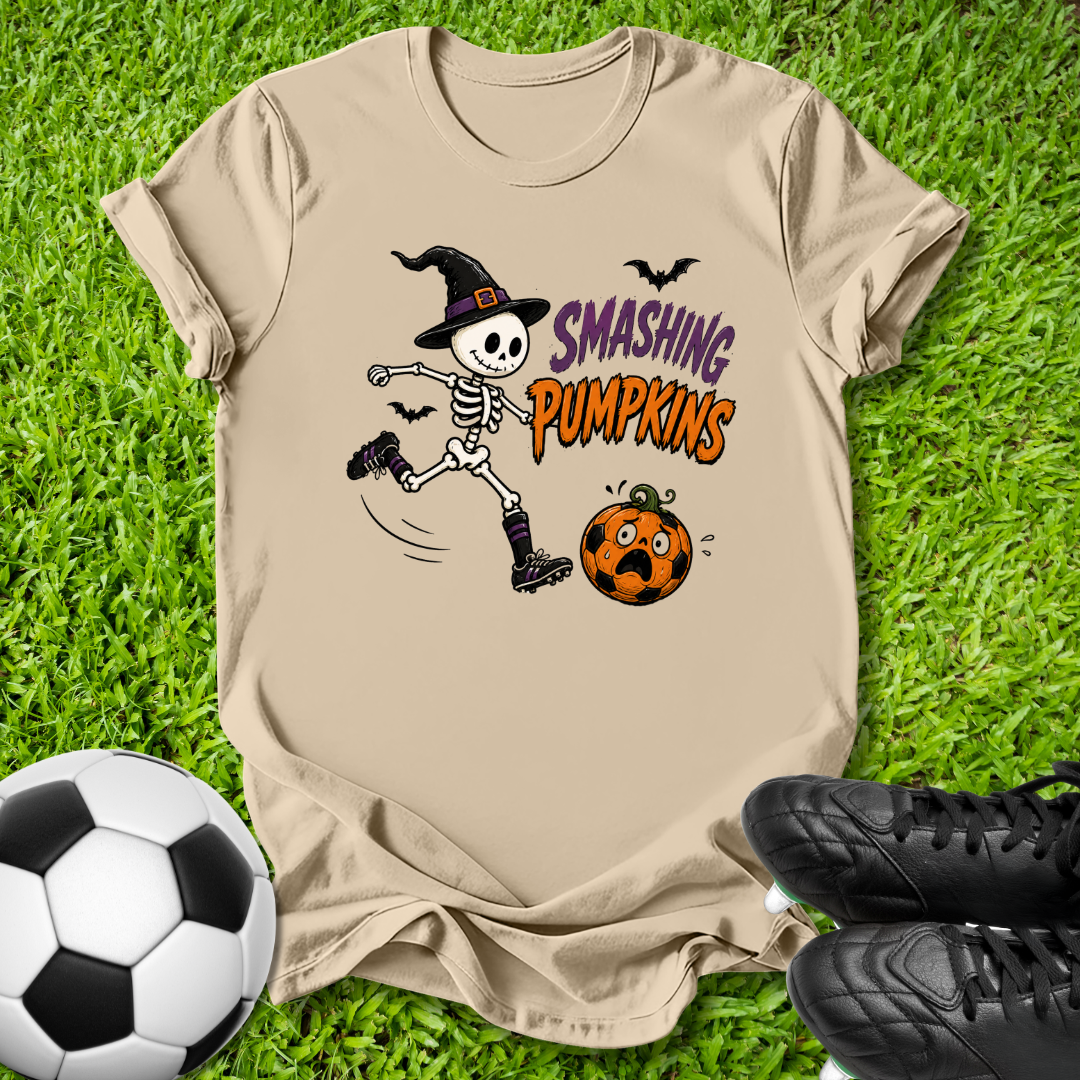A beige graphic t-shirt featuring a cartoon skeleton in a witch hat kicking a pumpkin with the text 'SMASHING PUMPKINS', laid flat on green grass with a soccer ball and black cleats as props.