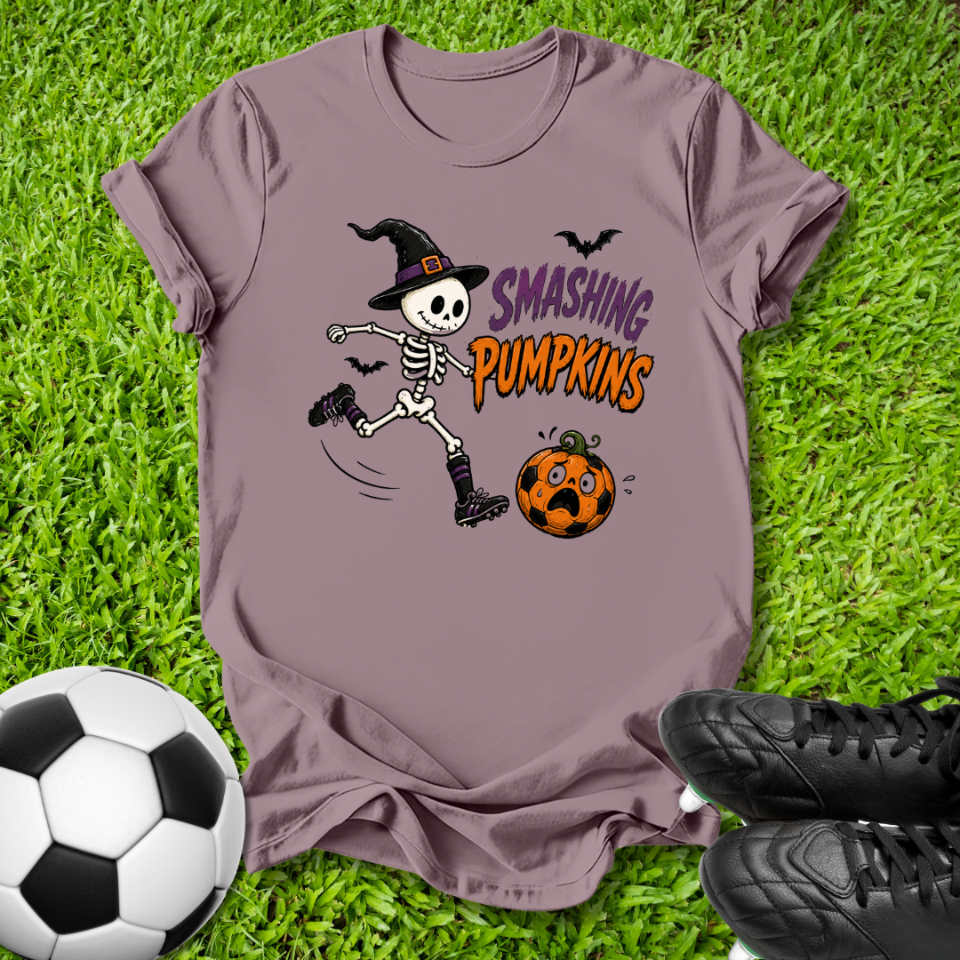 A mauve short-sleeve t-shirt laid flat on green grass, featuring a cartoon skeleton in a witch hat kicking a jack-o'-lantern with the text 'SMASHING PUMPKINS'. A soccer ball and black cleats are placed around it as props.