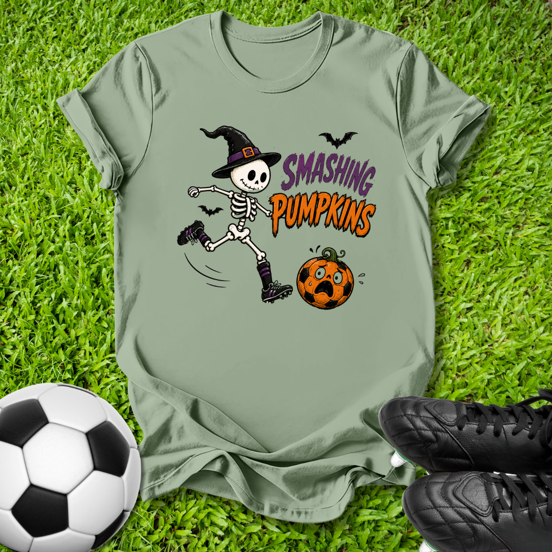 An olive green graphic t-shirt laid flat on a green grass background, styled with a soccer ball and black cleats. The t-shirt features a cartoon skeleton wearing a witch hat and kicking a jack-o'-lantern pumpkin, with the text 'SMASHING PUMPKINS' in purple and orange.