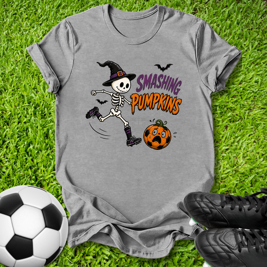 A heather gray graphic t-shirt featuring a cartoon skeleton kicking a pumpkin with the text 'SMASHING PUMPKINS', styled on green grass with a soccer ball and black cleats.