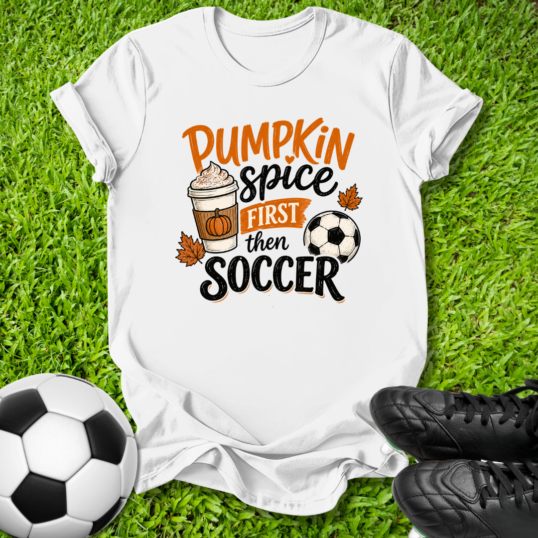 A white graphic t-shirt laid flat on green grass, featuring the text 'Pumpkin Spice First Then Soccer' with a coffee cup, pumpkin, and soccer ball illustration. A soccer ball and black cleats are placed around it as thematic props.