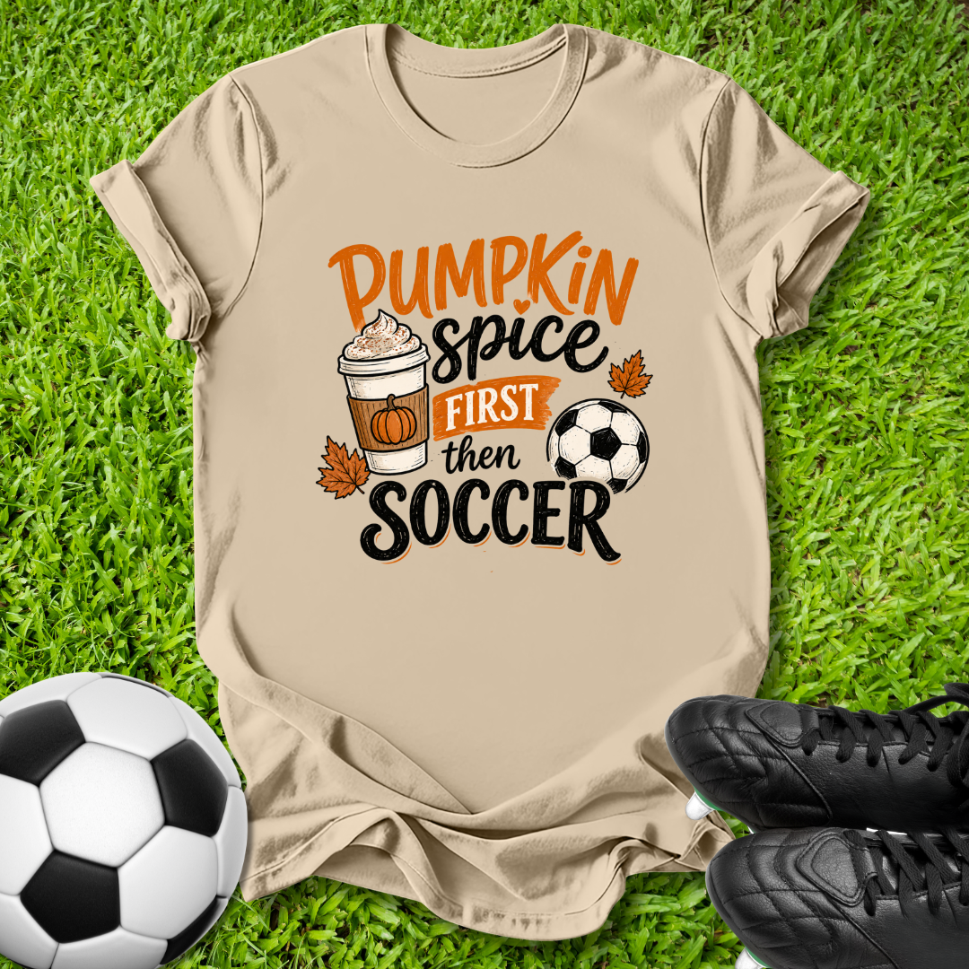 A beige graphic t-shirt laid flat on green grass, featuring the text 'Pumpkin Spice FIRST then SOCCER' with a coffee cup, pumpkin, leaves, and soccer ball illustration. A soccer ball and black cleats are placed around it as props.