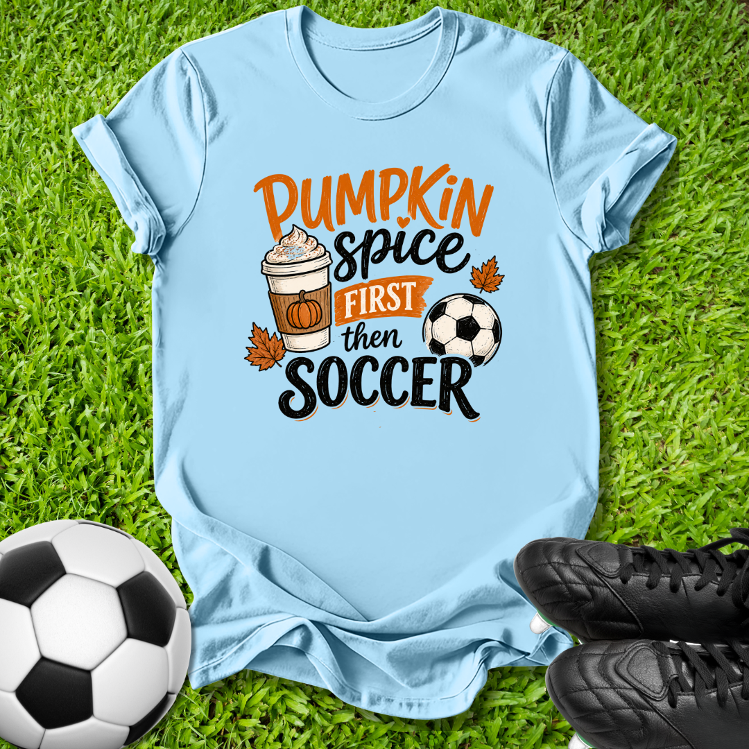 A light blue graphic t-shirt laid flat on green grass, featuring the text 'Pumpkin Spice First Then Soccer' with autumn-themed illustrations. A soccer ball and black cleats are placed around it as props.