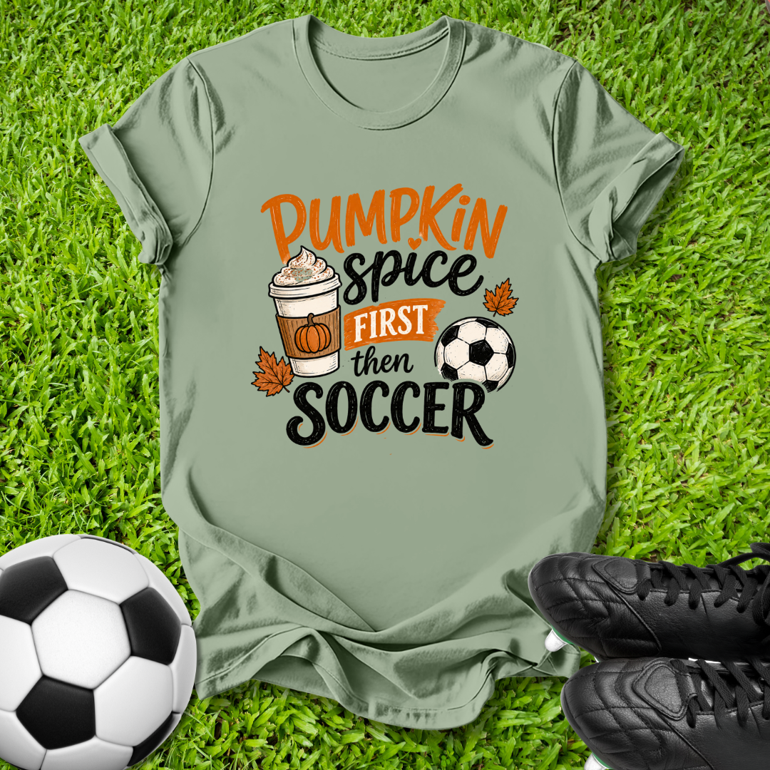An olive green graphic t-shirt laid flat on green grass, styled with soccer balls and black cleats. The shirt features a graphic that reads 'Pumpkin spice FIRST then SOCCER' in orange and black lettering, accompanied by illustrations of a pumpkin coffee cup, autumn leaves, and a soccer ball.