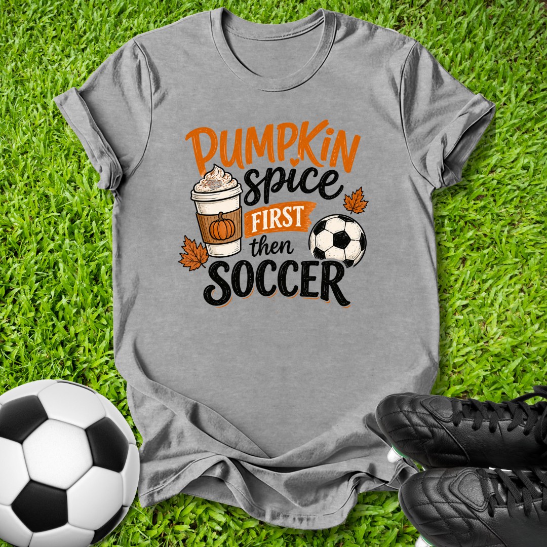 A heather gray graphic t-shirt laid flat on green grass, featuring the text 'Pumpkin Spice FIRST then SOCCER' with a coffee cup, pumpkin, autumn leaves, and soccer ball illustration. A soccer ball and black cleats are placed around it as props.