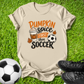 A beige graphic t-shirt laid flat on green grass, featuring the text 'Pumpkin Spice FIRST then SOCCER' with a coffee cup, pumpkin, and soccer ball illustration. A soccer ball and black cleats are placed around it as props.