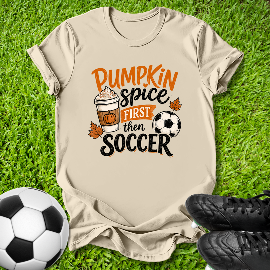 A beige graphic t-shirt laid flat on green grass, featuring the text 'Pumpkin Spice FIRST then SOCCER' with a coffee cup, pumpkin, and soccer ball illustration. A soccer ball and black cleats are placed around it as props.