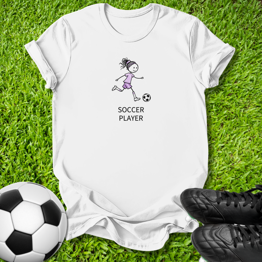 A white short-sleeve t-shirt laid flat on green grass, featuring a simple black line drawing of a girl playing soccer with the words 'SOCCER PLAYER' printed below. A soccer ball is visible at the bottom left and black cleats at the bottom right.