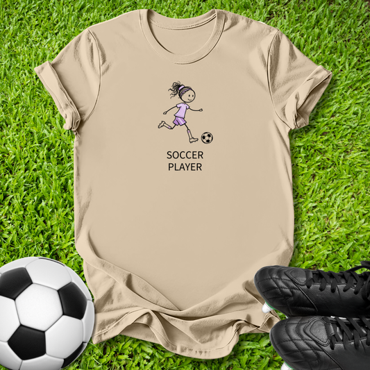 A beige graphic t-shirt featuring a cartoon illustration of a girl playing soccer with the text 'SOCCER PLAYER' underneath, laid flat on green grass with a soccer ball and black cleats as props.