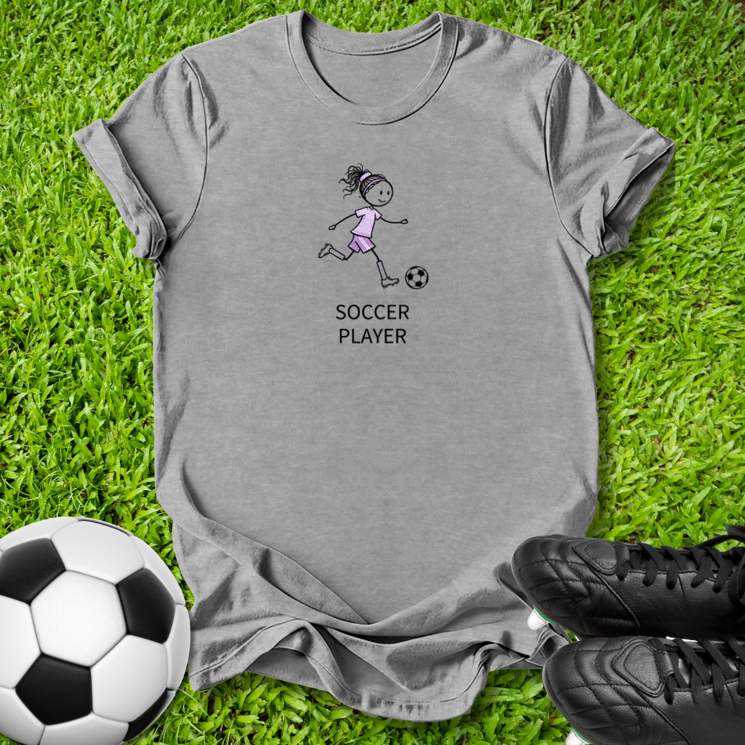 A heather gray short-sleeve t-shirt laid flat on green grass, featuring a cartoon girl soccer player graphic with the text 'SOCCER PLAYER'. A soccer ball and black cleats are styled around the shirt.