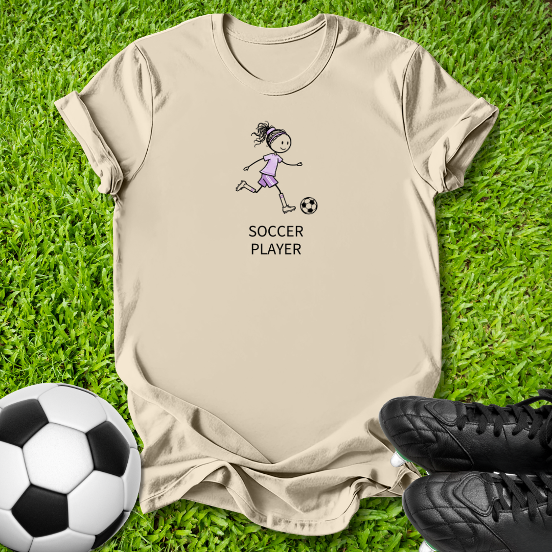 A beige graphic t-shirt laid flat on green grass, featuring a cartoon illustration of a girl playing soccer with the text 'SOCCER PLAYER' underneath. A soccer ball is visible in the bottom-left corner and a pair of black soccer cleats is in the bottom-right corner.