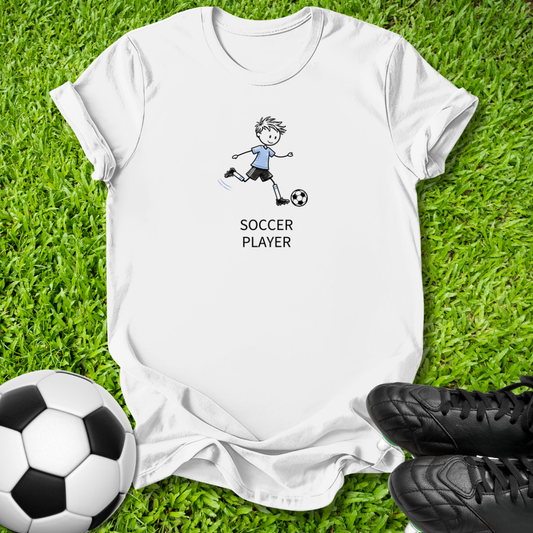 A white short-sleeve t-shirt laid flat on green grass, featuring a cartoon drawing of a soccer player kicking a ball with the text 'SOCCER PLAYER' underneath. A black-and-white soccer ball is in the bottom-left corner, and a pair of black soccer cleats is in the bottom-right corner.