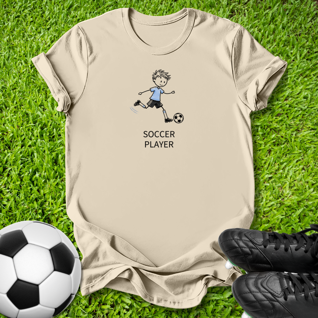 A beige short-sleeve t-shirt laid flat on green grass, featuring a cartoon graphic of a soccer player kicking a ball with the text 'SOCCER PLAYER' underneath. A black-and-white soccer ball is at the bottom left, and black cleats are at the bottom right.