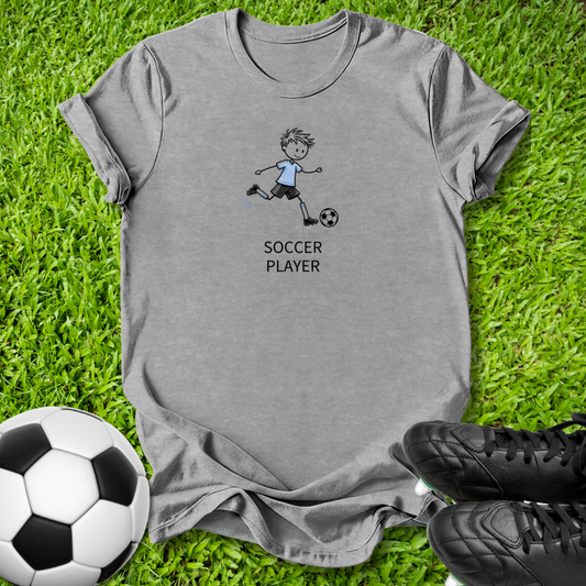 A heather gray short-sleeve t-shirt with a cartoon soccer player graphic and the text 'SOCCER PLAYER' printed on the front, laid flat on green grass with a soccer ball and black cleats nearby.