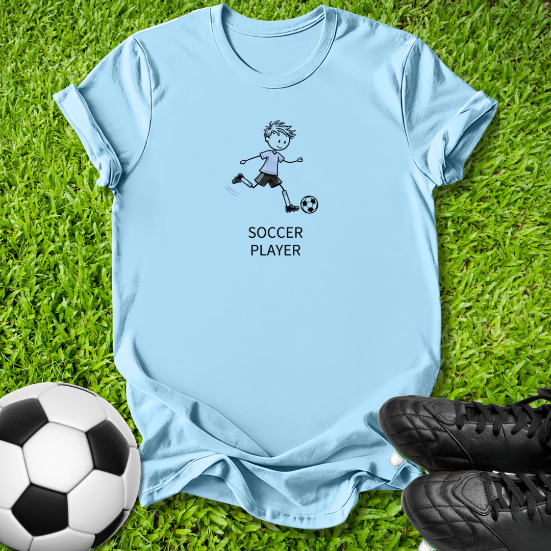 A light blue short-sleeve t-shirt laid flat on green grass, featuring a cartoon soccer player graphic and the words 'SOCCER PLAYER' printed on the front. A soccer ball is visible at the bottom left, and black cleats are at the bottom right.