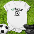 A white short-sleeve t-shirt laid flat on green grass, featuring the word 'unlucky' in green and black text above a black-and-white soccer ball graphic. A soccer ball and black cleats are placed around it as props.