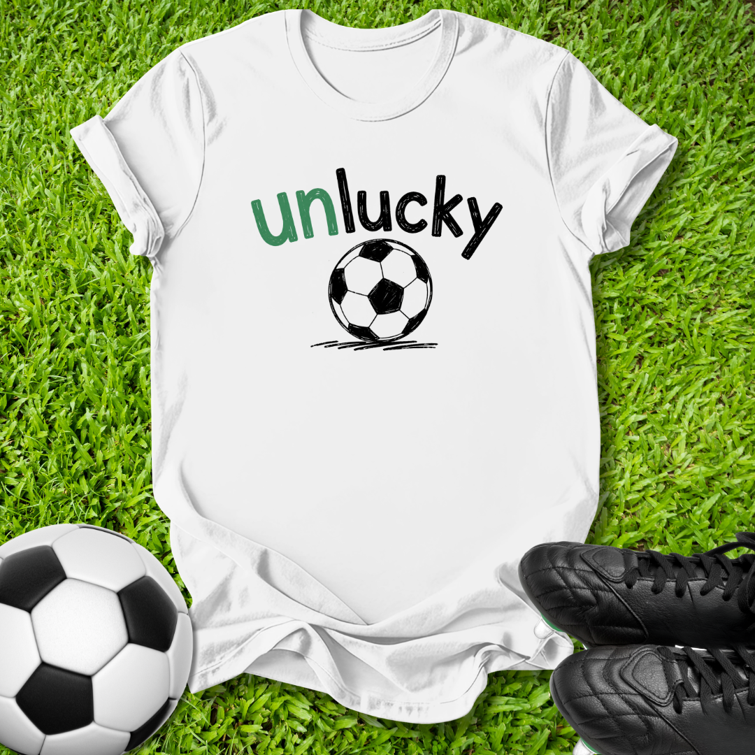 A white short-sleeve t-shirt laid flat on green grass, featuring the word 'unlucky' in green and black text above a black-and-white soccer ball graphic. A soccer ball and black cleats are placed around it as props.