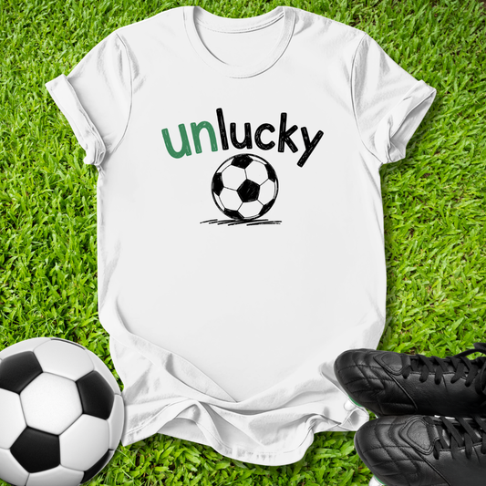 A white short-sleeve t-shirt laid flat on green grass, featuring the word 'unlucky' in green and black text above a black-and-white soccer ball graphic. A soccer ball and black cleats are placed around it as props.