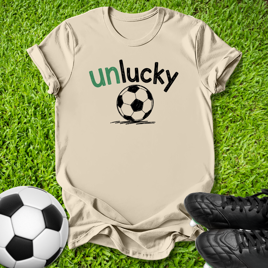 A beige short-sleeve t-shirt laid flat on green grass, featuring the text 'unlucky' in green and black above a black-and-white soccer ball graphic. A real soccer ball and black cleats are placed around it as props.