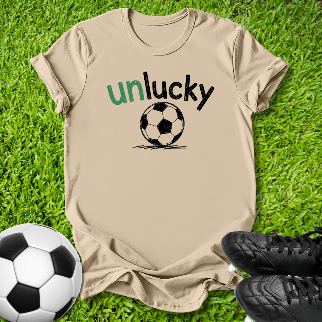 A beige graphic t-shirt laid flat on green grass, featuring the text 'unlucky' in green and black above a black-and-white soccer ball. A soccer ball and black cleats are placed around it as props.