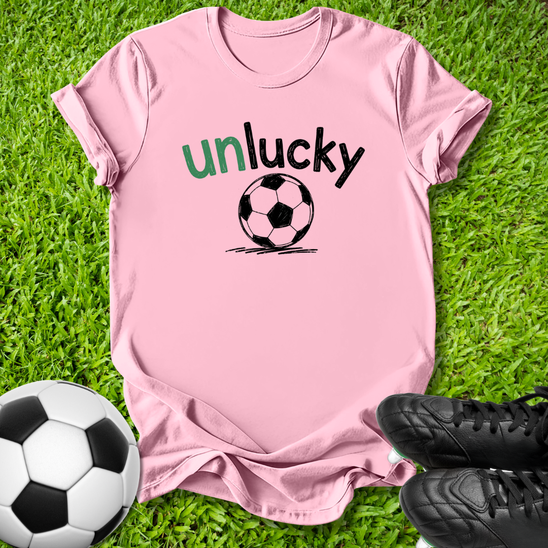 A light pink short-sleeve t-shirt with a graphic that reads 'unlucky' in green and black text above a black and white soccer ball, laid flat on green grass with a soccer ball and black cleats as props.