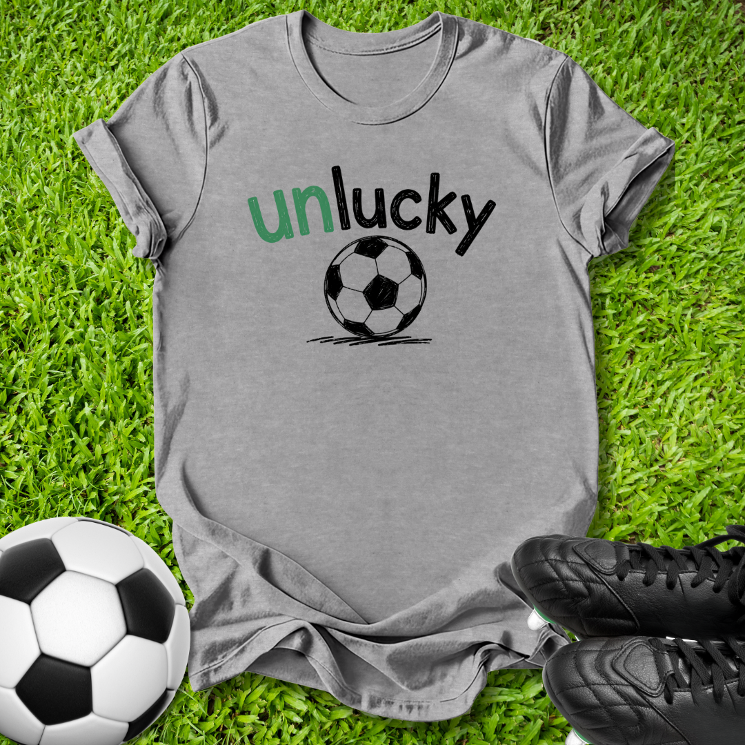 A heather gray t-shirt with a graphic that reads 'unlucky' in green and black text above a black and white soccer ball, laid flat on green grass with a soccer ball and black cleats as props.