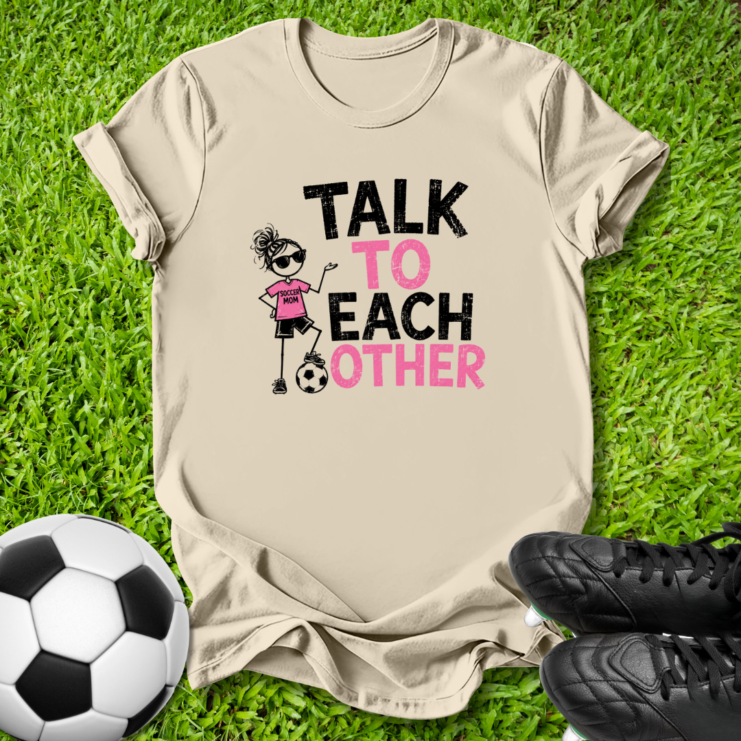 A beige graphic t-shirt laid flat on green grass, featuring a cartoon soccer girl and the text 'TALK TO EACH OTHER' in black and pink. A soccer ball is on the left and black cleats are on the right.