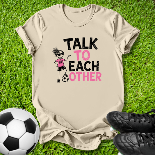 A beige graphic t-shirt laid flat on green grass, featuring a cartoon soccer girl and the text 'TALK TO EACH OTHER' in black and pink. A soccer ball is on the left and black cleats are on the right.