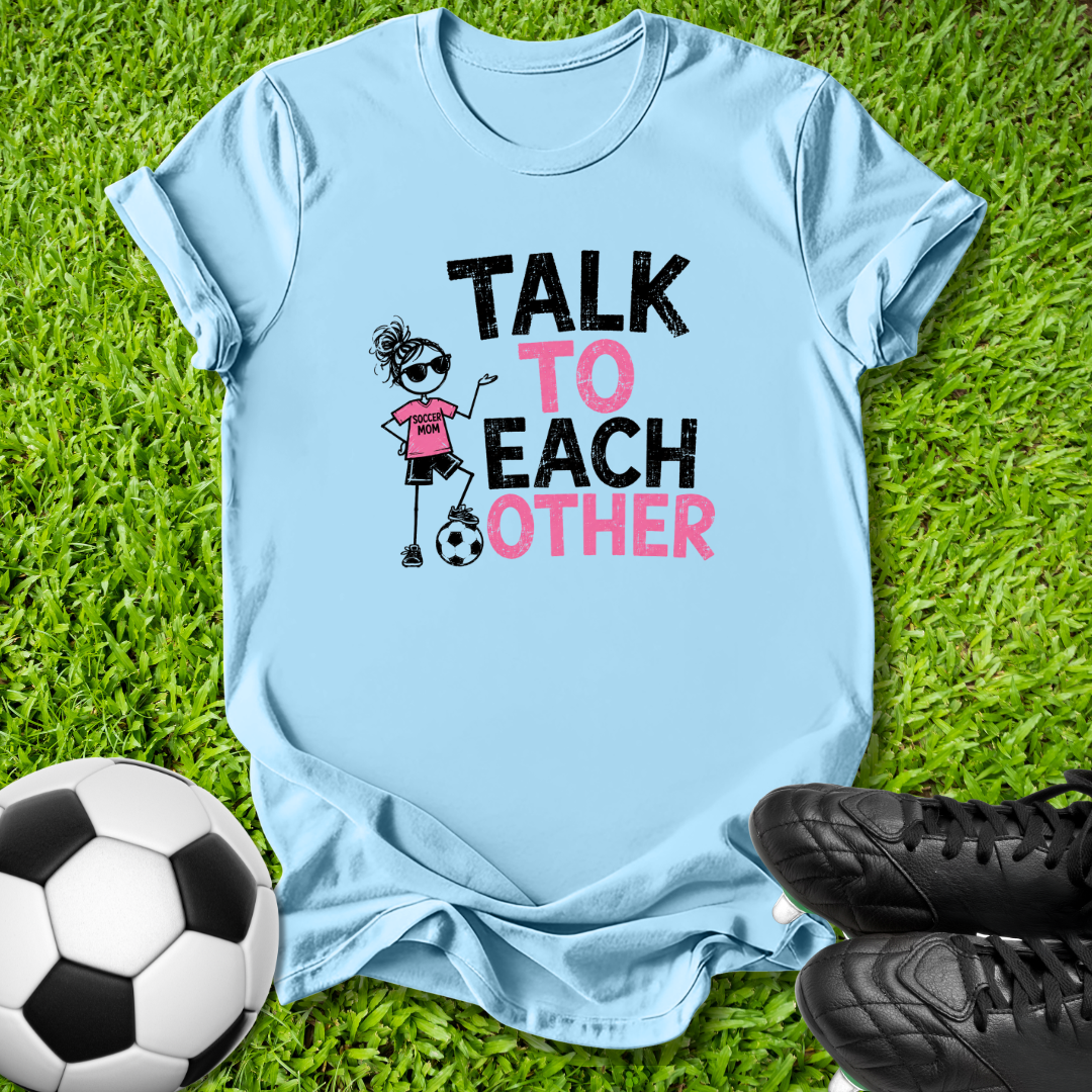 A light blue graphic t-shirt laid flat on green grass, featuring the text 'TALK TO EACH OTHER' in black and pink with a cartoon soccer-themed illustration. A soccer ball and black cleats are placed around it as props.