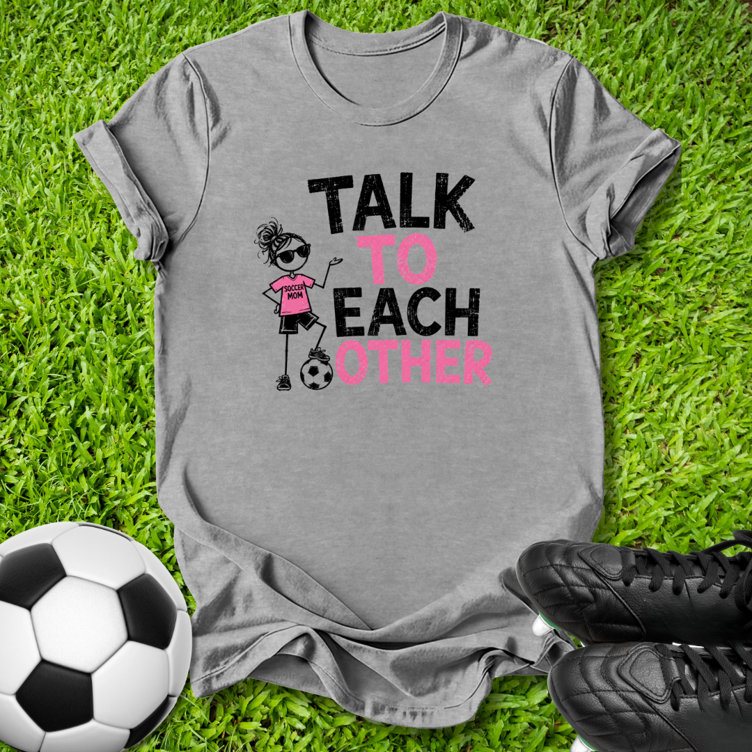 A heather gray graphic t-shirt laid flat on green grass, featuring a cartoon girl kicking a soccer ball with the text 'TALK TO EACH OTHER' in black and pink. A soccer ball and black cleats are placed around it as props.