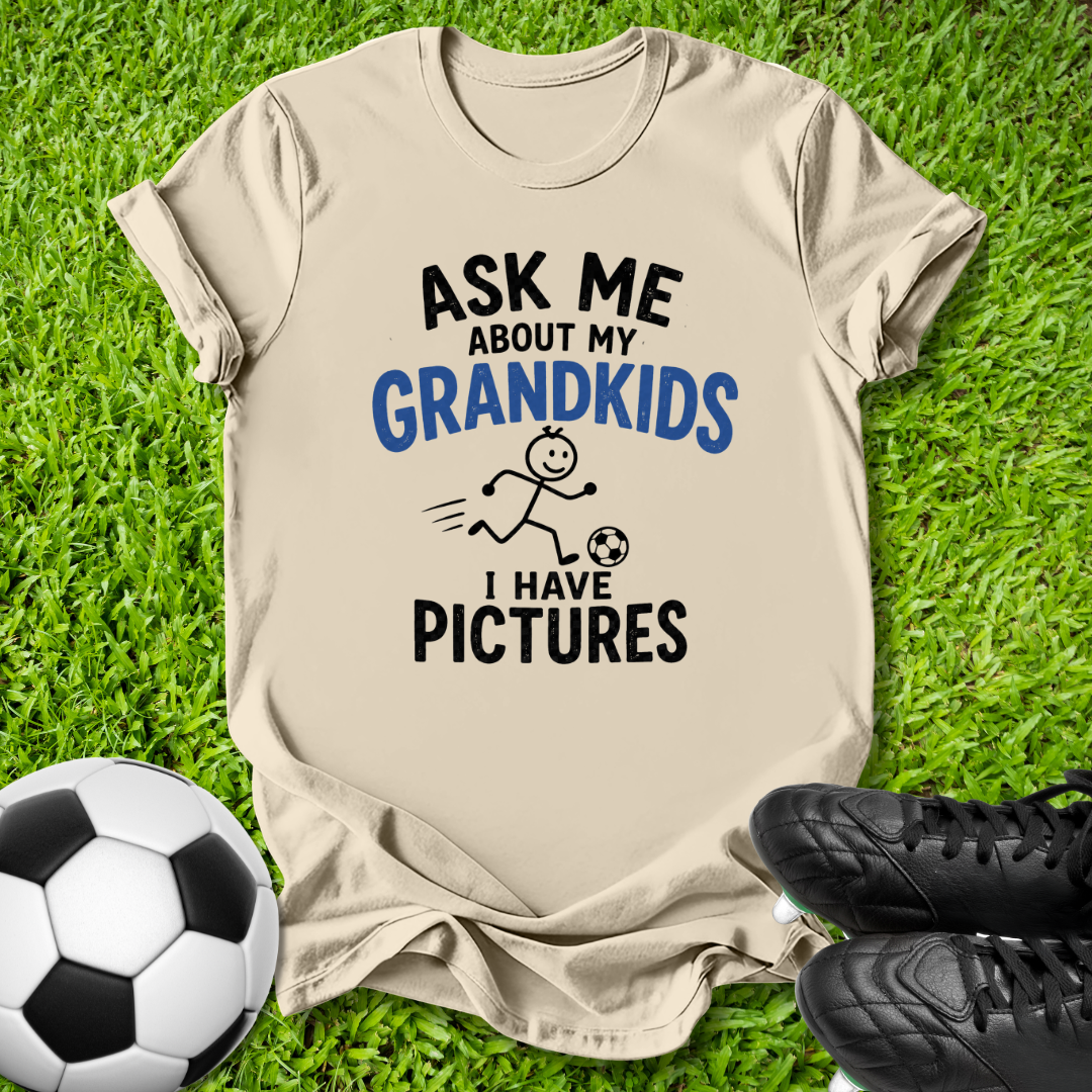 A beige graphic t-shirt laid flat on green grass, featuring the text 'ASK ME ABOUT MY GRANDKIDS I HAVE PICTURES' in black and blue, with a small stick figure soccer graphic. A soccer ball and black cleats are placed around it as props.