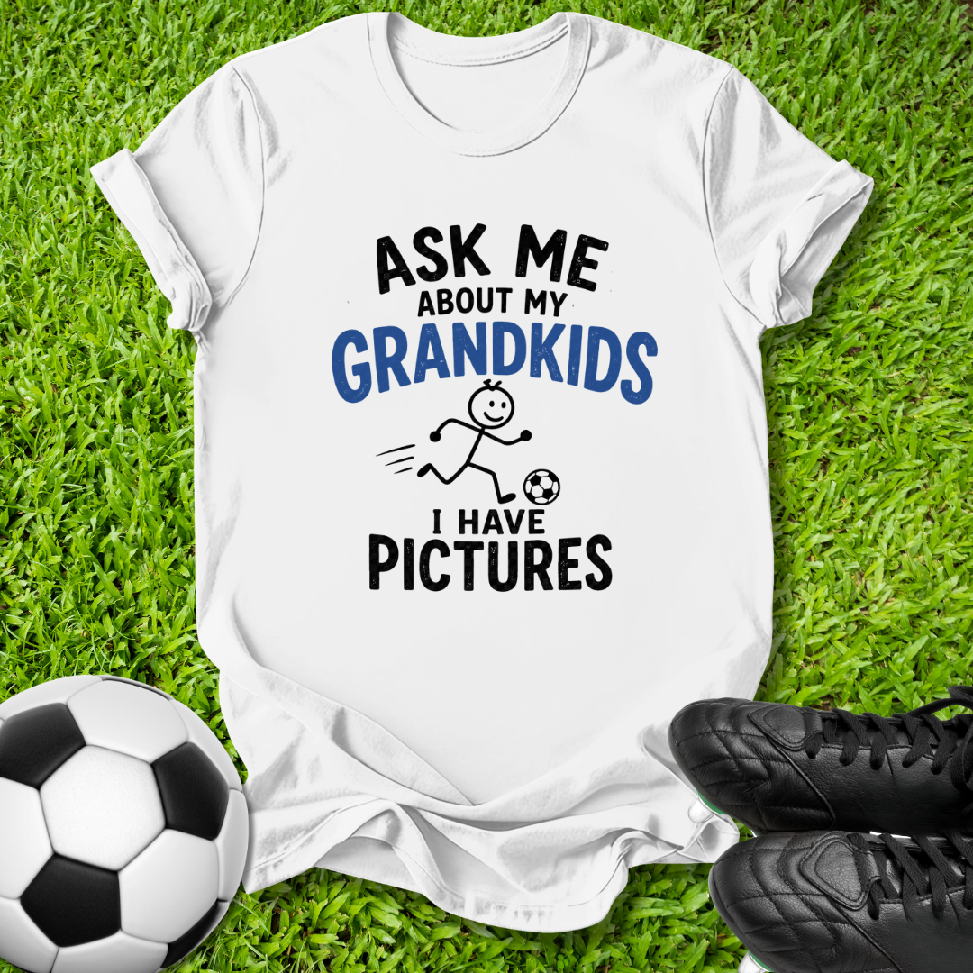 A white graphic t-shirt laid flat on green grass, featuring the text 'ASK ME ABOUT MY GRANDKIDS I HAVE PICTURES' in black and blue lettering with a stick figure kicking a soccer ball. A soccer ball and black cleats are placed around the shirt as props.
