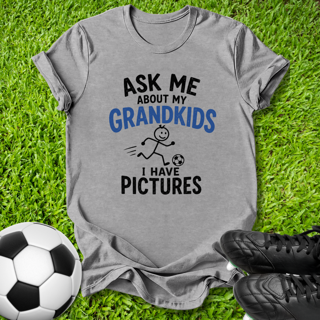A heather gray graphic t-shirt laid flat on green grass, featuring the text 'ASK ME ABOUT MY GRANDKIDS I HAVE PICTURES' in black and blue, with a small stick figure kicking a soccer ball. A soccer ball and black cleats are placed around it as props.