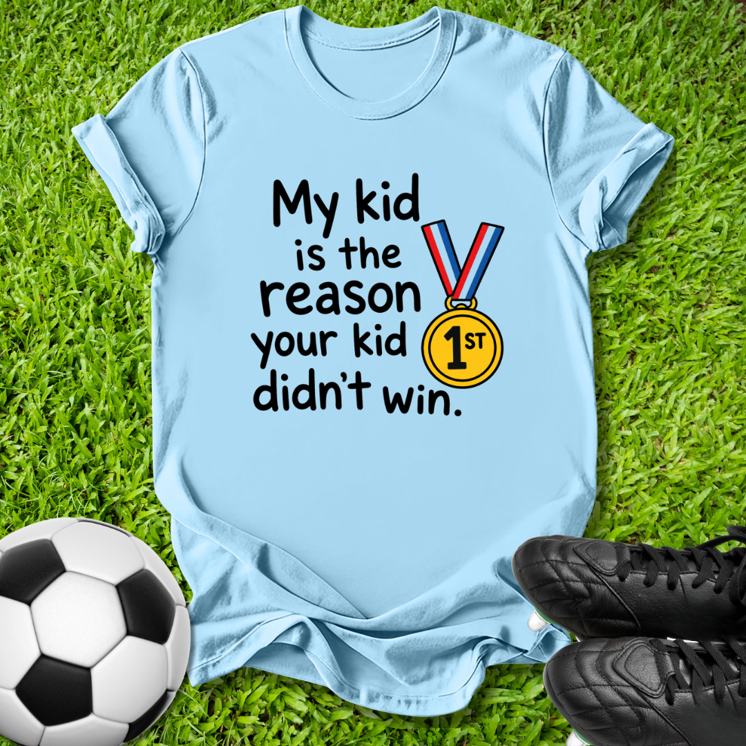 A light blue graphic t-shirt laid flat on green grass, featuring the text 'My kid is the reason your kid didn't win.' alongside a gold medal graphic.