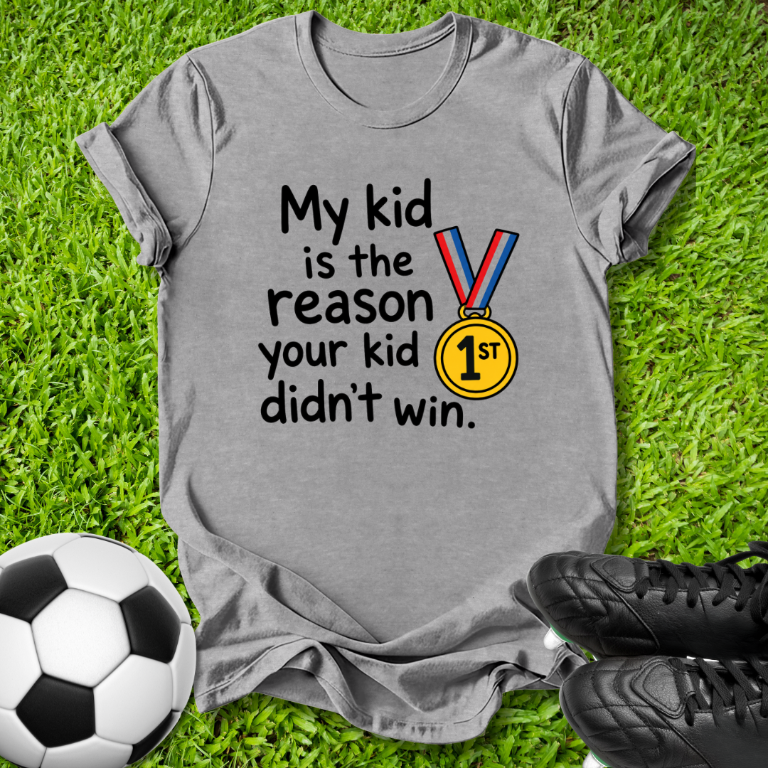 A heather gray short-sleeve t-shirt laid flat on green grass, featuring the printed text 'My kid is the reason your kid didn't win.' alongside a gold medal graphic with a blue and red ribbon.