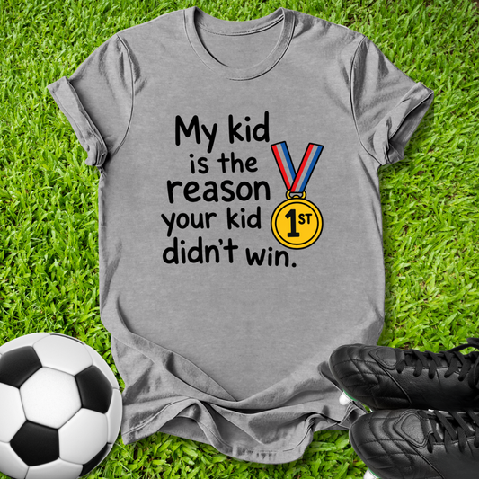 A heather gray short-sleeve t-shirt laid flat on green grass, featuring the printed text 'My kid is the reason your kid didn't win.' alongside a gold medal graphic with a blue and red ribbon.