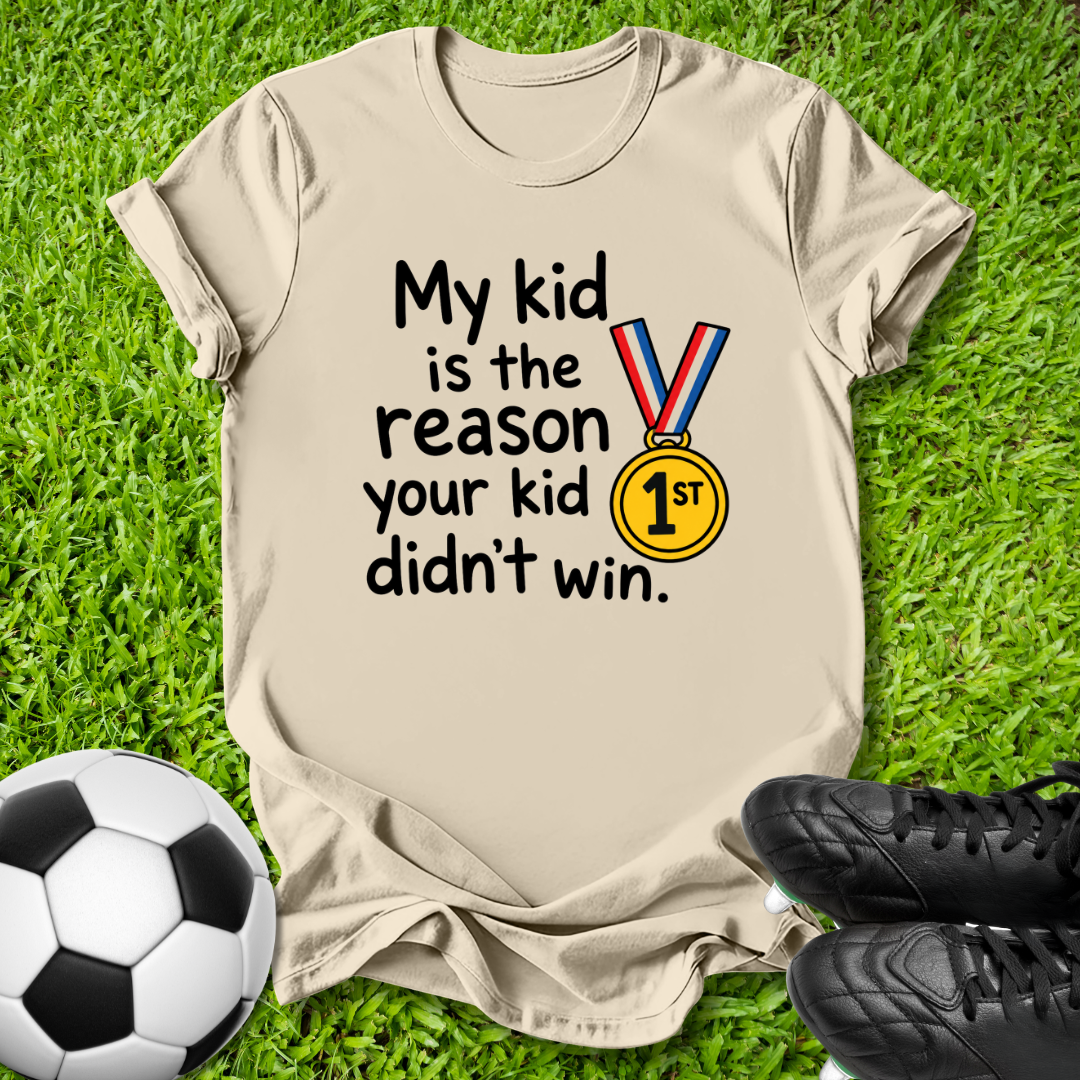 A beige graphic t-shirt laid flat on green grass, featuring the text 'My kid is the reason your kid didn't win.' alongside a yellow gold medal graphic.