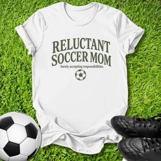 A white short-sleeve T-shirt laid flat on green grass, printed with the words 'RELUCTANT SOCCER MOM' and 'barely accepting responsibilities.' plus a soccer ball graphic.