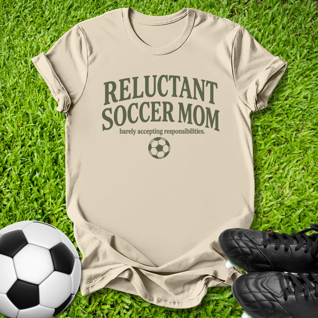 A beige graphic t-shirt laid flat on green grass, featuring the text 'RELUCTANT SOCCER MOM' and 'barely accepting responsibilities.' with a soccer ball icon.