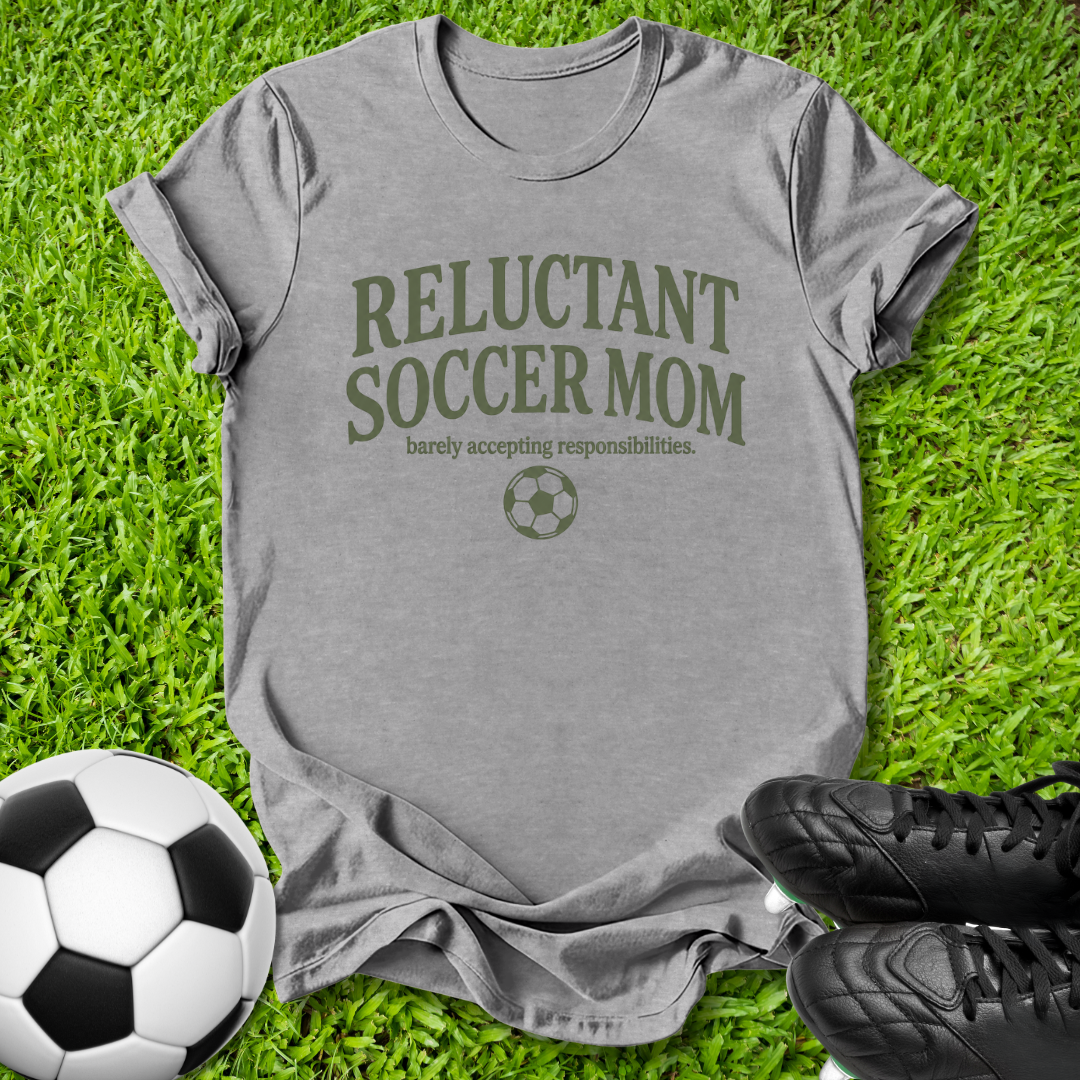 A heather gray t-shirt with 'RELUCTANT SOCCER MOM' printed in olive green on the front, styled on a green grass background with a soccer ball and black cleats.