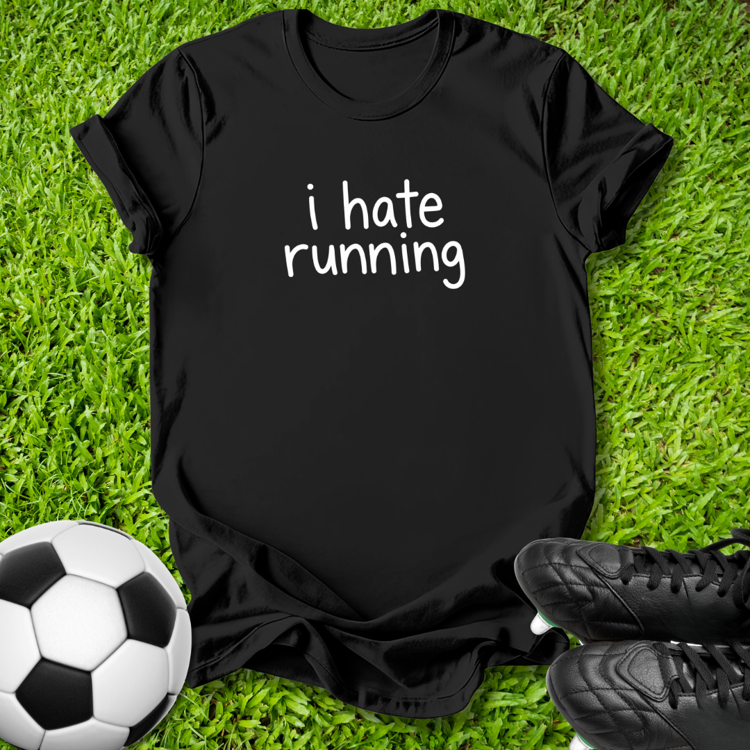 A black short-sleeve t-shirt with the phrase 'i hate running' printed in white lowercase letters, laid flat on green grass with a soccer ball and black cleats as props.