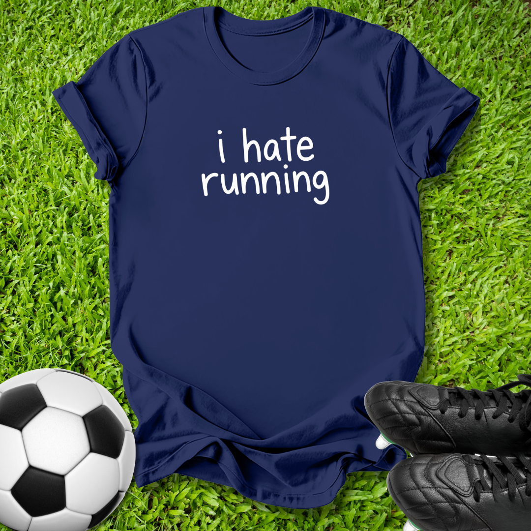 A navy blue t-shirt with the text 'i hate running' printed in white on the front, laid flat on green grass with a soccer ball and black cleats as props.