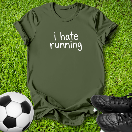 An olive green t-shirt with the text 'i hate running' printed in white on the chest, laid flat on a green grass background with a soccer ball and black cleats as props.