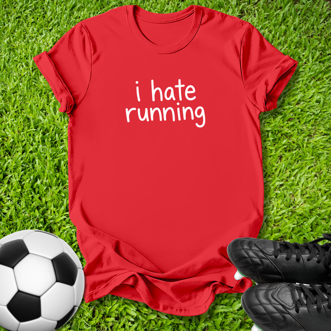 A red short-sleeve t-shirt with the white text 'i hate running' printed on the chest, laid flat on green grass with a soccer ball and black cleats as props.