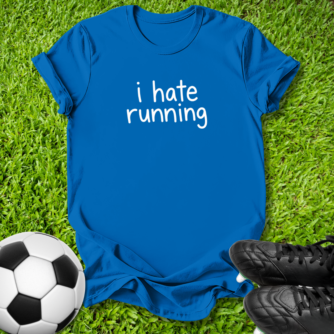 A royal blue short-sleeve t-shirt with the phrase 'i hate running' printed in white lowercase letters across the chest, laid flat on green grass with a soccer ball and black cleats as props.