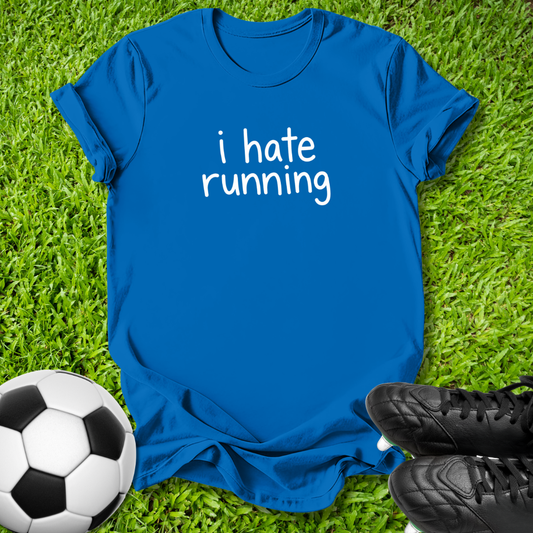 A royal blue short-sleeve t-shirt with the phrase 'i hate running' printed in white lowercase letters across the chest, laid flat on green grass with a soccer ball and black cleats as props.