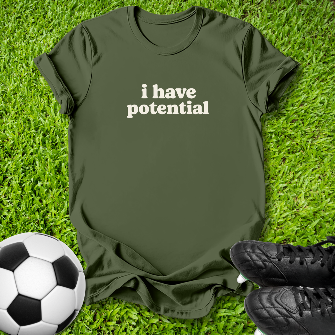 An olive green t-shirt with the text 'i have potential' printed in white on the chest, laid flat on a green grass background with a soccer ball and black cleats as props.